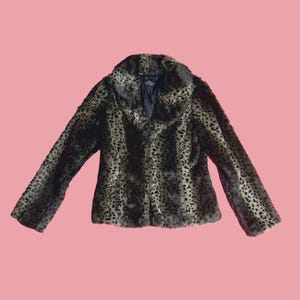 May include: A faux fur jacket with a leopard print pattern. The jacket has a wide collar and long sleeves. The background is a solid pink color. The jacket is a neutral color with black and brown spots.