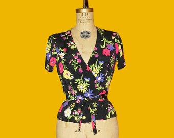 May include: A black wrap top with a floral print featuring pink, yellow, and blue flowers. The top has short sleeves and a V-neckline. The top is tied at the waist with a black sash.