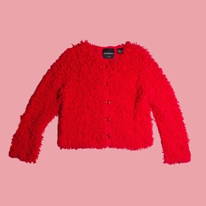 May include: A bright red long-sleeved cardigan with a textured, shaggy knit surface. The garment features a round neckline, a front button closure with matching red buttons, and a black interior label with the text Michael Simon.