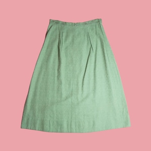 May include: A light mint green A-line skirt featuring a textured fabric with a subtle speckled pattern. The skirt has a high waistband with a series of small, matching buttons along the top edge and two vertical pleats at the front waistline.