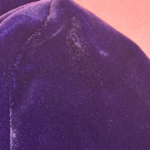 May include: Close-up of a section of a deep purple velvet fabric. The fabric has a soft, plush texture and a slight sheen. The image shows the corner of a garment, with a seam visible.