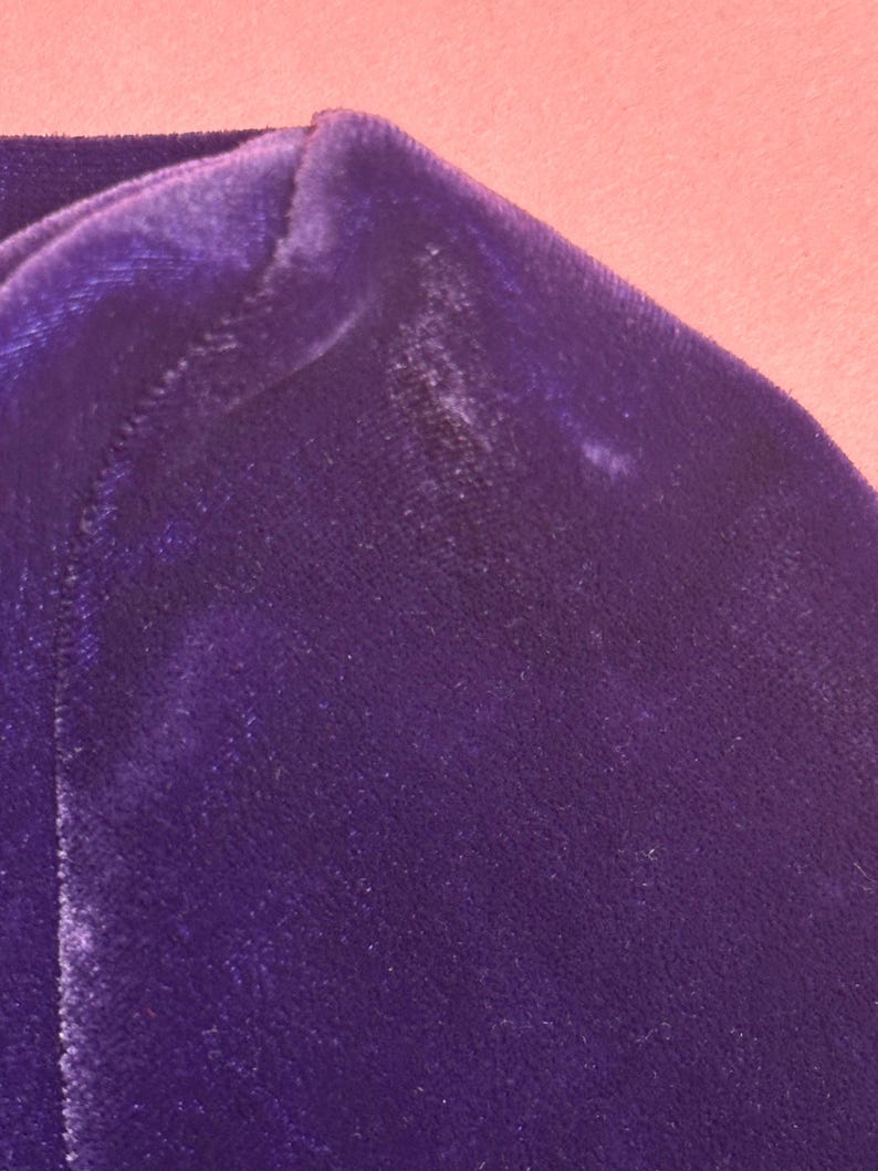 May include: Close-up of a section of a deep purple velvet fabric. The fabric has a soft, plush texture and a slight sheen. The image shows the corner of a garment, with a seam visible.