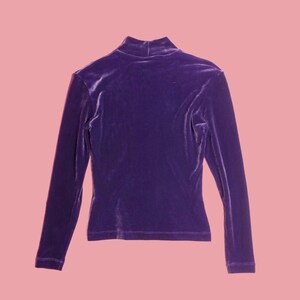 May include: A long-sleeved, purple velvet top with a mock neck. The top is a solid color and has a fitted silhouette. The fabric appears soft and has a slight sheen. The background is a solid pink.