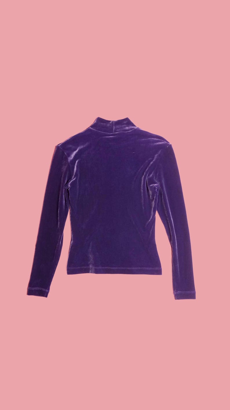 May include: A long-sleeved, purple velvet top with a mock neck. The top is a solid color and has a fitted silhouette. The fabric appears soft and has a slight sheen. The background is a solid pink.