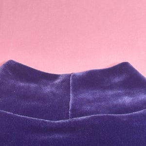 May include: Close-up of a deep purple velvet mock neck top. The fabric has a soft, plush texture. The background is a solid light pink color.