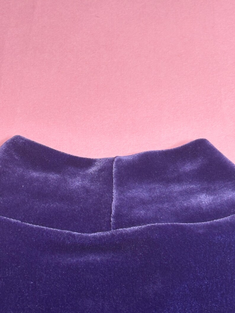 May include: Close-up of a deep purple velvet mock neck top. The fabric has a soft, plush texture. The background is a solid light pink color.