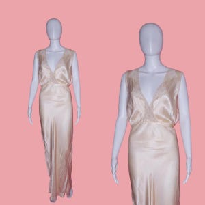 May include: A cream-colored satin gown with a V-neckline and delicate lace trim. The sleeveless dress has a flowing, floor-length skirt and is displayed on a mannequin against a pink background. The gown's design is simple yet elegant.