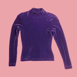 May include: A long-sleeved, purple velvet top with a mock neck. The top is a solid color and has a fitted silhouette. The garment is displayed on a pink background.