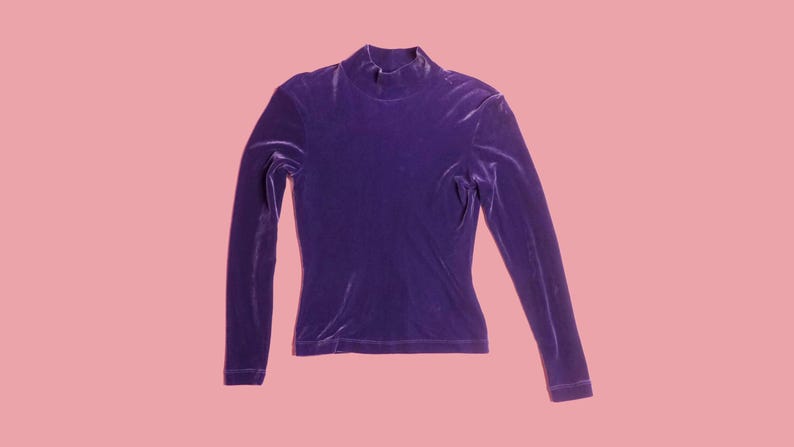 May include: A long-sleeved, purple velvet top with a mock neck. The top is a solid color and has a fitted silhouette. The garment is displayed on a pink background.