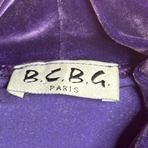 May include: Close-up of a purple velvet hooded top. The label reads "B.C.B.G. PARIS" in black lettering on a white tag. The fabric has a soft, textured appearance.