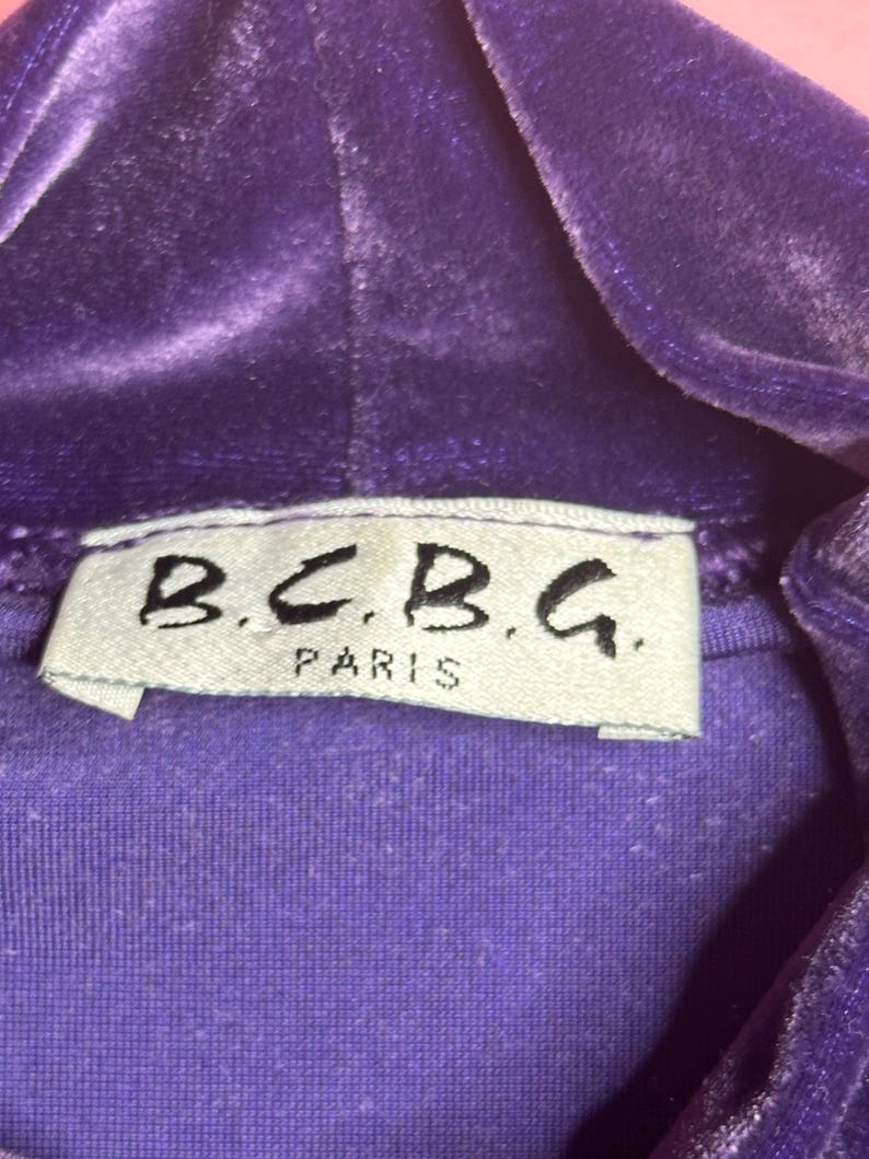 May include: Close-up of a purple velvet hooded top. The label reads "B.C.B.G. PARIS" in black lettering on a white tag. The fabric has a soft, textured appearance.