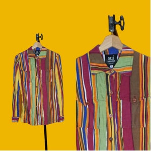 May include: A long-sleeved button-down shirt with vertical stripes in various colors, including yellow, green, red, and brown. The shirt has two chest pockets and is hanging on a wooden hanger. The label reads "sui anna sui".