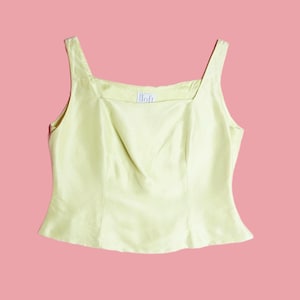 May include: A pale yellow square-neck tank top with a slightly gathered bodice. The top has a fitted silhouette and thin straps. The word "lloft" is printed on the front. The background is a solid pink.