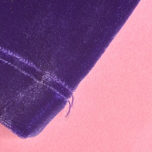 May include: Close-up of a section of deep purple velvet fabric. The fabric has a soft, plush texture and is slightly angled. The edge of the fabric is visible, with a few loose threads. The background is a solid light pink.