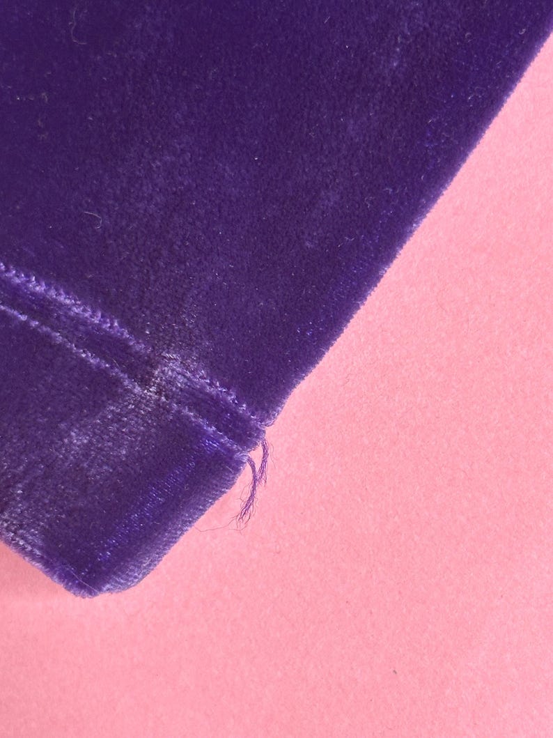 May include: Close-up of a section of deep purple velvet fabric. The fabric has a soft, plush texture and is slightly angled. The edge of the fabric is visible, with a few loose threads. The background is a solid light pink.