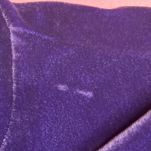 May include: Close-up of a section of deep purple velvet fabric. The fabric is folded, revealing its texture and sheen. The background is a soft pink. There are two small, lighter-colored spots on the velvet.