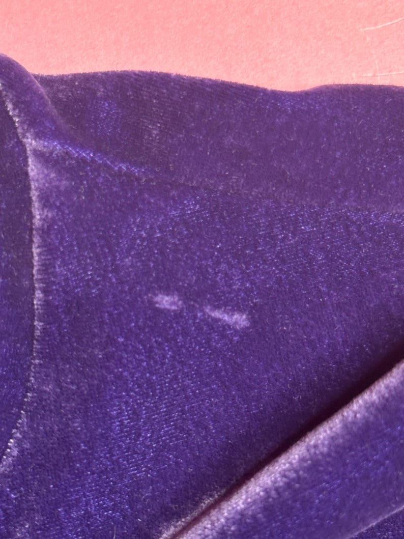 May include: Close-up of a section of deep purple velvet fabric. The fabric is folded, revealing its texture and sheen. The background is a soft pink. There are two small, lighter-colored spots on the velvet.
