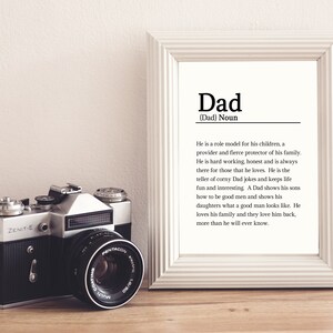 May include: A white framed print with the word "Dad" in black text. The print defines the word "Dad" as a noun and describes the qualities of a father. A vintage camera is in the foreground.