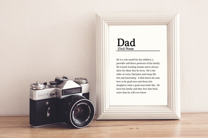 May include: A white framed print with the word "Dad" in black text. The print defines the word "Dad" as a noun and describes the qualities of a father. A vintage camera is in the foreground.