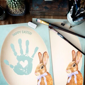 Puede incluir: Dos tarjetas imprimibles con un conejo y una huella de mano en un círculo azul. El texto "¡Feliz Pascua!" está en la parte superior de la tarjeta.