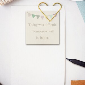 May include: A white card with a gold heart-shaped paper clip holding it to a white sheet of paper. The card has a colorful banner design at the top and the text "Today was difficult. Tomorrow will be better."