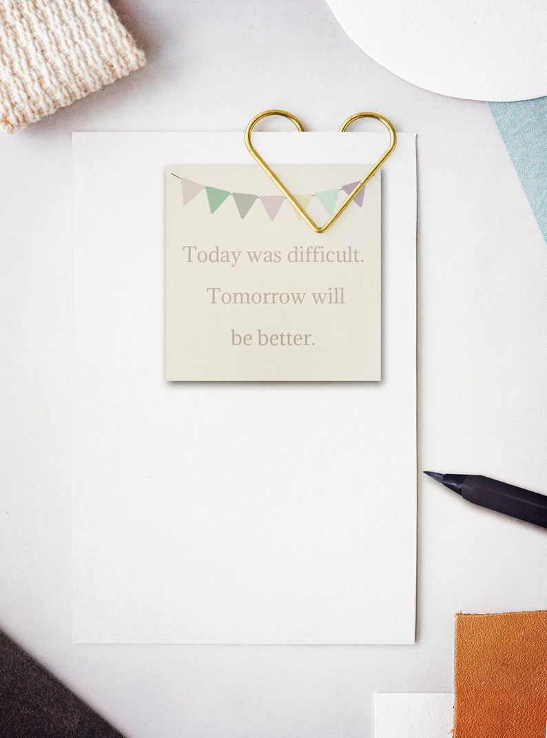 May include: A white card with a gold heart-shaped paper clip holding it to a white sheet of paper. The card has a colorful banner design at the top and the text "Today was difficult. Tomorrow will be better."