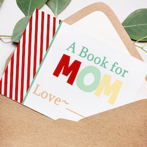 May include: A greeting card with the text "A Book for MOM" in red, green, and yellow, inside a brown envelope. The card has a red and white striped pattern on the left side. Eucalyptus leaves are in the background.