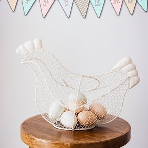 May include: White wire chicken-shaped egg holder with a banner that reads "Happy Easter" above it. The holder is filled with white and brown eggs and sits on a wooden stool.