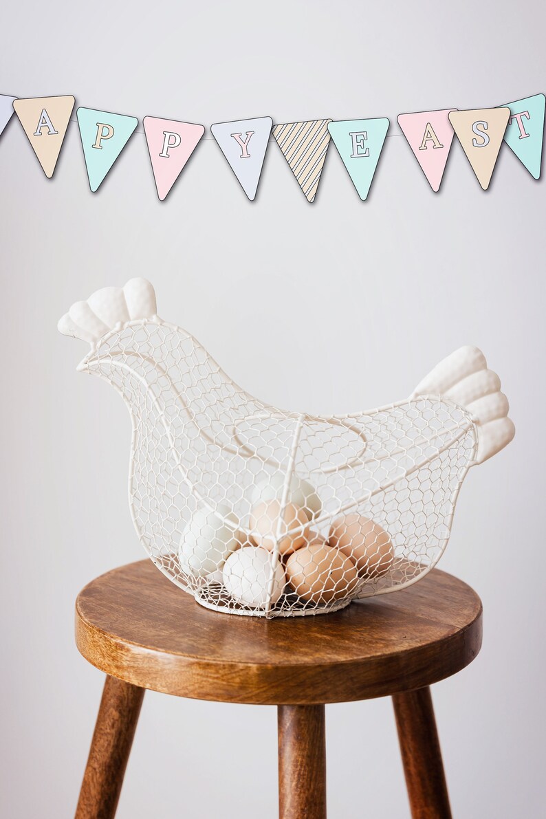 May include: White wire chicken-shaped egg holder with a banner that reads "Happy Easter" above it. The holder is filled with white and brown eggs and sits on a wooden stool.