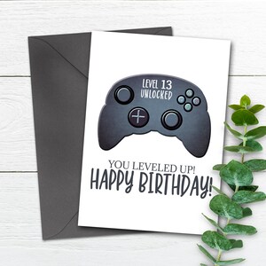 May include: A white card with a black and gray video game controller graphic. The text "Level 13 Unlocked" is above the controller and the text "You Leveled Up! Happy Birthday!" is below the controller.