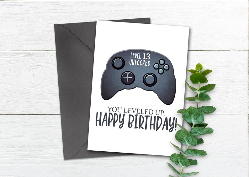 May include: A white card with a black and gray video game controller graphic. The text "Level 13 Unlocked" is above the controller and the text "You Leveled Up! Happy Birthday!" is below the controller.