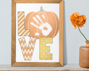 Può includere: Una stampa incorniciata con la parola "LOVE" in lettere decorative. La "L" ha strisce diagonali, la "V" ha un motivo a zig-zag e la "E" ha pois bianchi. Una zucca con un'impronta della mano è sullo sfondo. Un vaso con un fiore arancione è a destra.