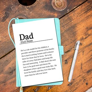May include: A light blue notebook with a white page that defines the word "Dad" as a noun. The definition describes a dad as a role model, provider, protector, and a hard-working, honest person who is always there for those he loves. The notebook is sitting on a wooden surface.