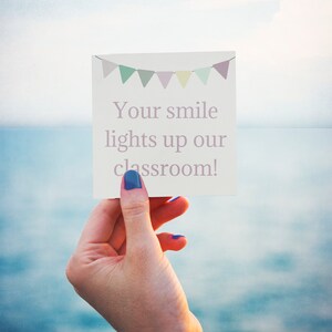 May include: A white card with a pink banner and the text "Your smile lights up our classroom!" held by a hand with blue nail polish.