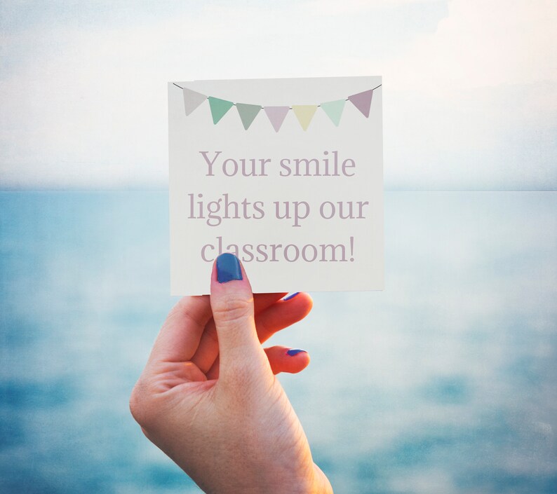 May include: A white card with a pink banner and the text "Your smile lights up our classroom!" held by a hand with blue nail polish.
