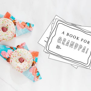 May include: A stack of blank white pages with a black border and the text "A BOOK FOR GRANDPA! BY..." on the top page. Two white donuts with pink frosting and colorful sprinkles are on top of blue and pink floral napkins.
