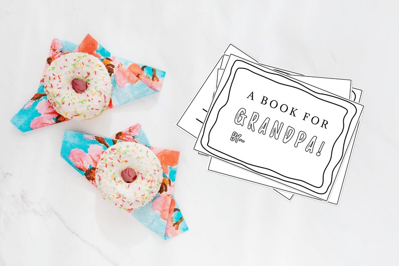 May include: A stack of blank white pages with a black border and the text "A BOOK FOR GRANDPA! BY..." on the top page. Two white donuts with pink frosting and colorful sprinkles are on top of blue and pink floral napkins.