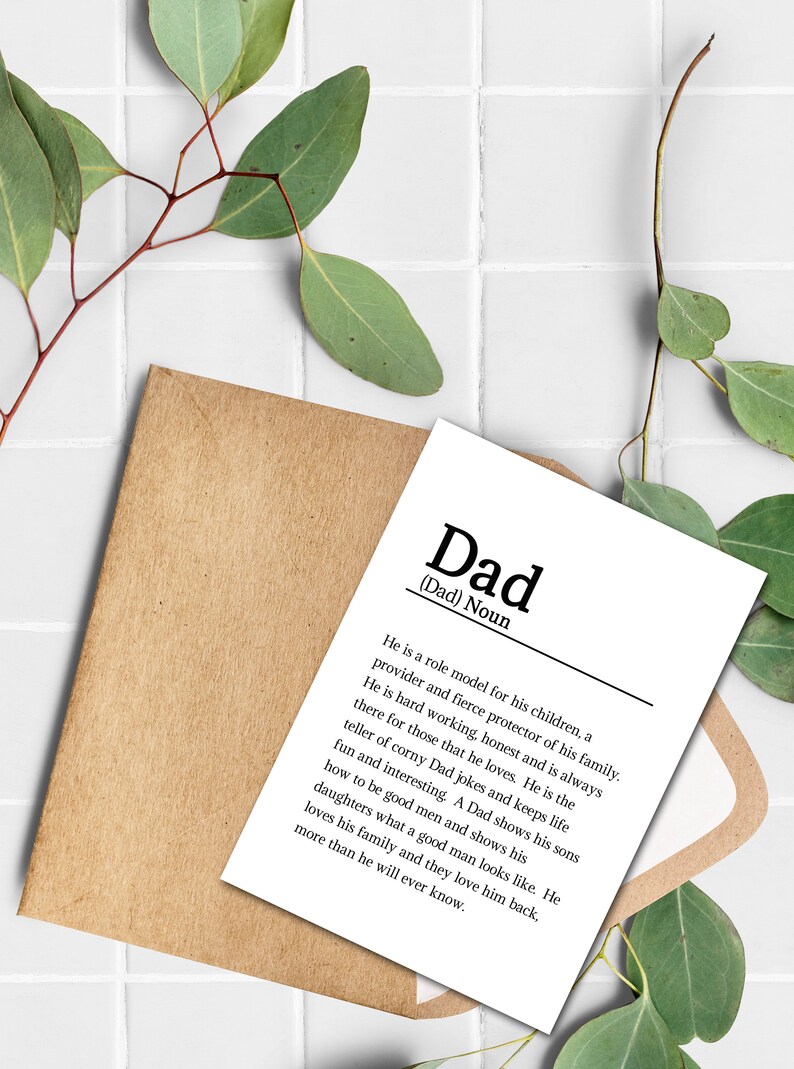 May include: A white card with black text defining the word "Dad" as a role model, provider, and protector of his family. The card is on a brown envelope with green leaves on a white tiled background.
