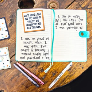 May include: A notebook with a turquoise cover and lined pages. The notebook is open and has a note card on the left page that says "Write about a time you felt proud of yourself and explain why you felt that way." The right page has handwritten text that says "I am so happy that my coach saw all the hard work I was putting in!" and "I was so proud of myself when I was given the award in Hockey. I worked really hard and practiced a lot."