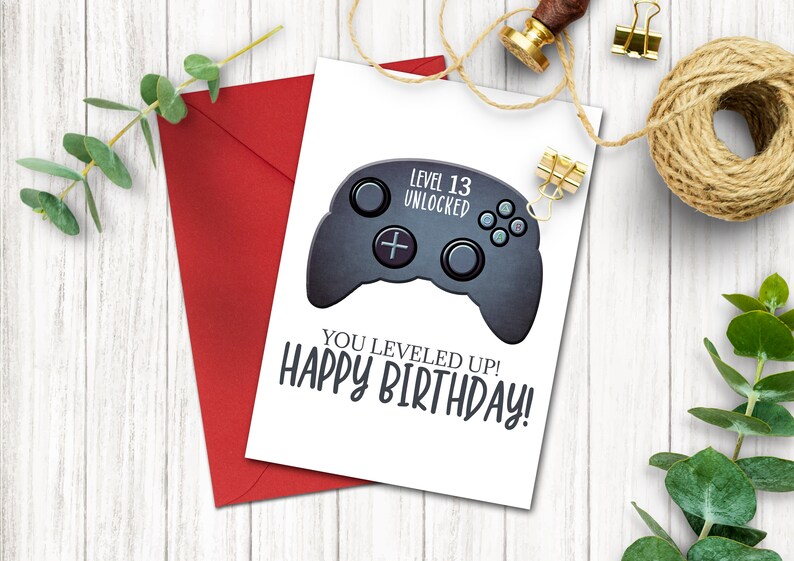 May include: A white card with a black and gray video game controller graphic. The text on the card reads "Level 13 Unlocked" and "You Leveled Up! Happy Birthday!"