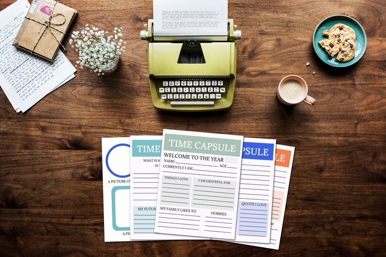 A table view with the 5 printable time capsule pages spread out on the table top. A typewriter, vase of flowers, plate of cookies and mug of coffee, some papers and a gift sit nearby.