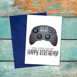 May include: A white card with a black and gray video game controller graphic. The text on the card reads "Level 13 Unlocked" and "You Leveled Up! Happy Birthday!"
