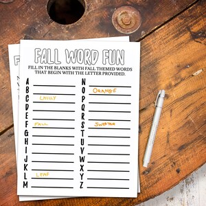 May include: A black and white "Fall Word Fun" worksheet with fall-themed words to fill in. The words "Chilly", "Fall", "Leaf", "Orange", and "Sweater" are already filled in. A silver pen rests on the wooden surface.
