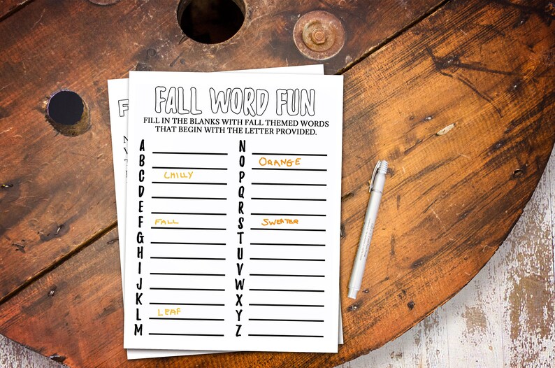 May include: A black and white "Fall Word Fun" worksheet with fall-themed words to fill in. The words "Chilly", "Fall", "Leaf", "Orange", and "Sweater" are already filled in. A silver pen rests on the wooden surface.