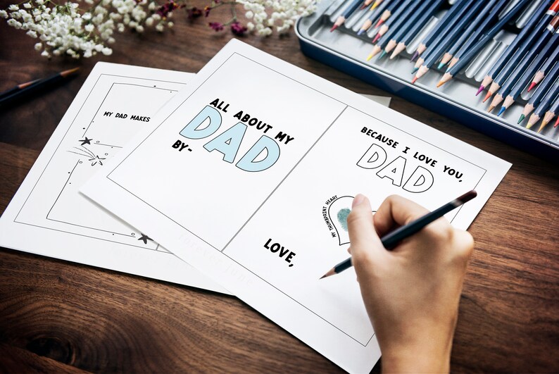 May include: A blank card with the text "All About My Dad" and "Because I Love You, Dad" with a heart shape for a fingerprint. The card is on a wooden table with a box of colored pencils.
