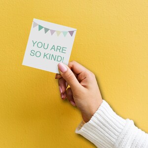 May include: A white card with a string of colorful flags at the top and the text "YOU ARE SO KIND!" in green letters. A hand is holding the card against a yellow background.