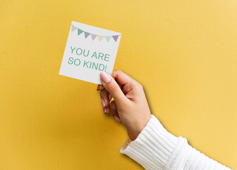May include: A white card with a string of colorful flags at the top and the text "YOU ARE SO KIND!" in green letters. A hand is holding the card against a yellow background.