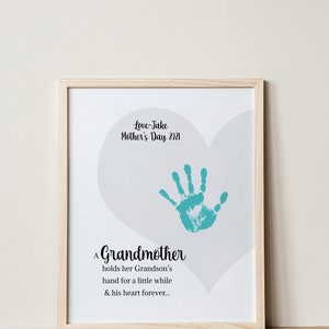 May include: A framed print with a light gray heart shape and a teal handprint inside. The text reads "Love-Jake Mother's Day 2021" and "A Grandmother holds her Grandson's hand for a little while & his heart forever..."