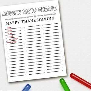 May include: A white paper with the text "AUTUMN WORD CREATE" and "HAPPY THANKSGIVING". Several words are written on the paper, with some highlighted in red. Three colored markers, blue, green, and red, are at the bottom.