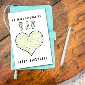 May include: A white card with the text "My heart belongs to Dad" in black lettering. The word "Dad" is in large, colorful letters. A yellow and blue heart with a black outline is below the text. The text "Happy Birthday!" is at the bottom of the card.